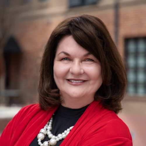 A person smiling outdoors, wearing a red jacket and pearl necklace with a blurred background of a brick building.