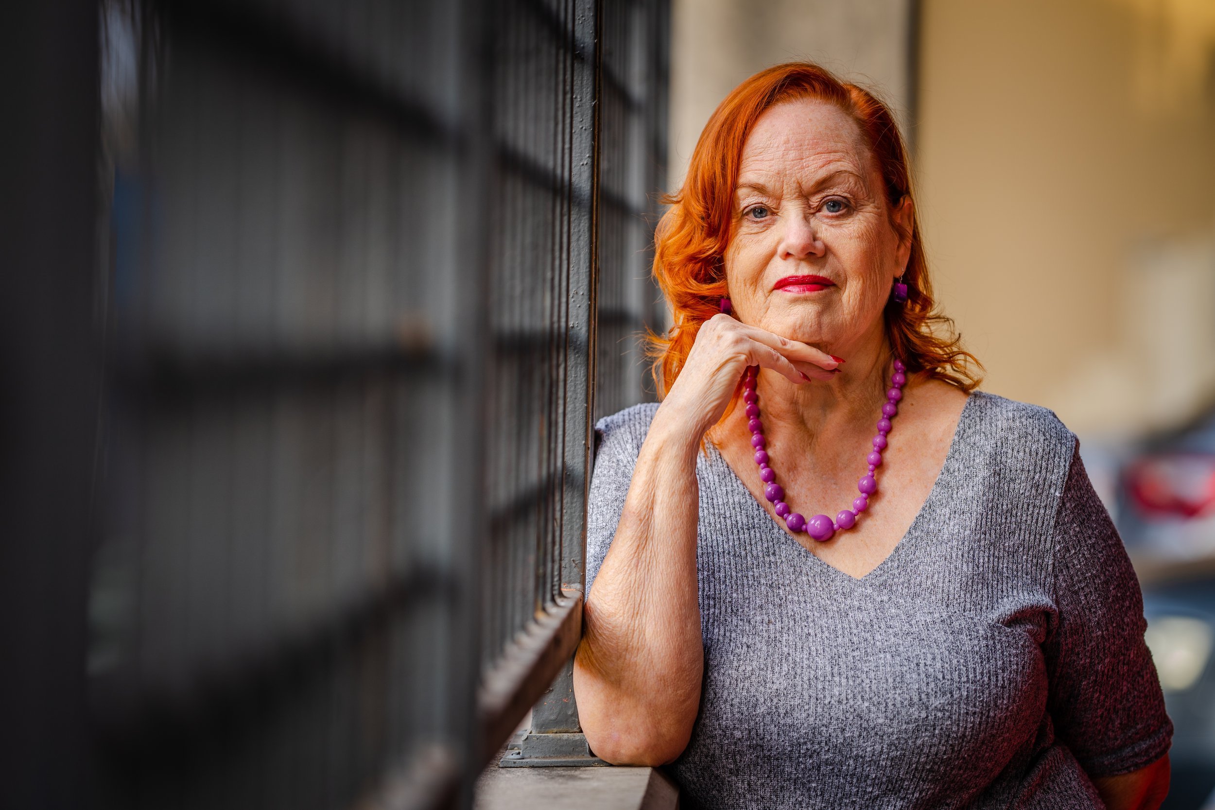 Elderly woman with red hair and purple necklace standing by a fence, smiling.