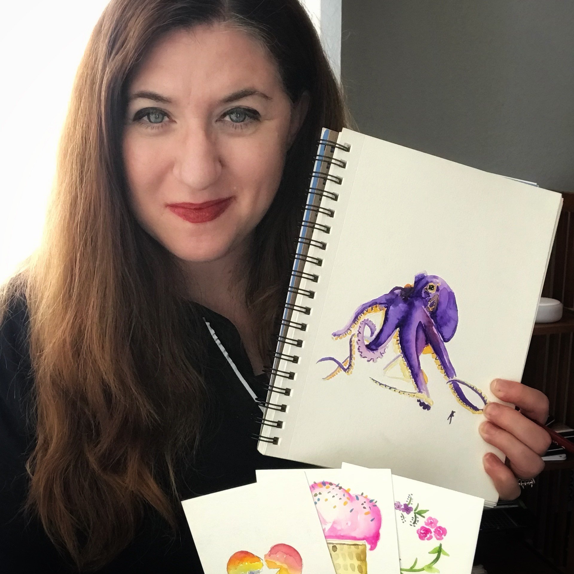 Artist Amy Pagan holding a wire-bound notebook to display a vibrant watercolor of an octopus in purples and yellows.