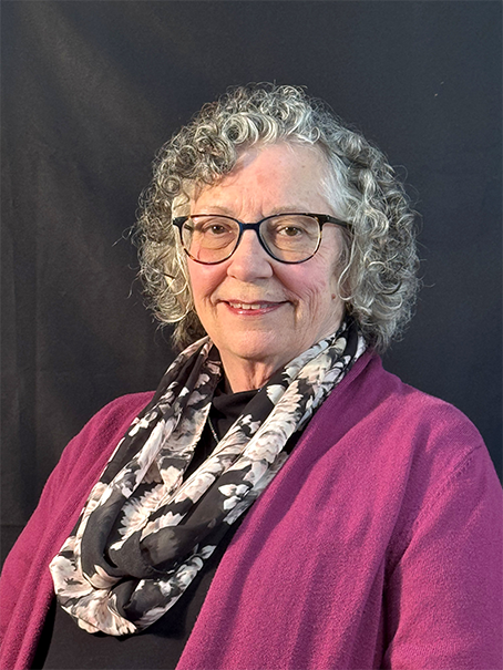 A smiling older woman with curly hair, wearing glasses and a floral scarf, against a dark background.